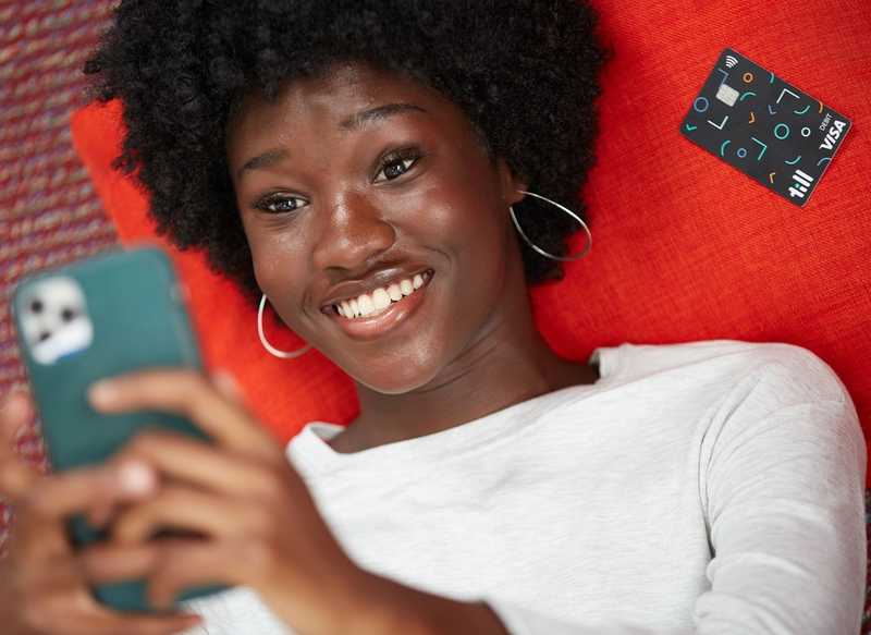 Teenager smiling while lying on the bed next to a Till debit card and looking at their Till app