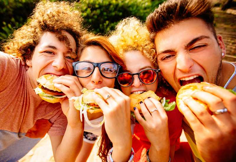 Four teens, two boys and two girls, biting into their hamburgers while taking a group selfie