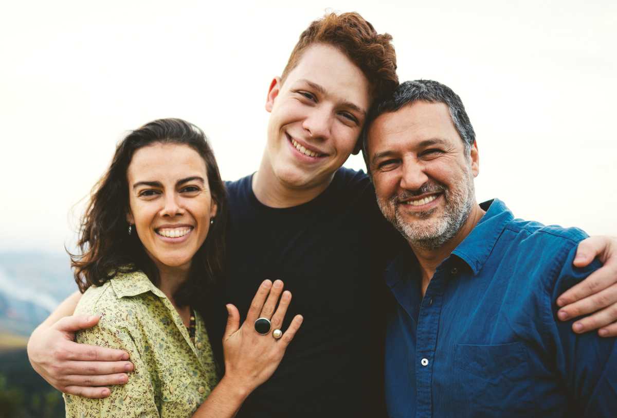 A family posing for the camera, the teenage son in the middle has his arms around his mom and dad on either side of him.